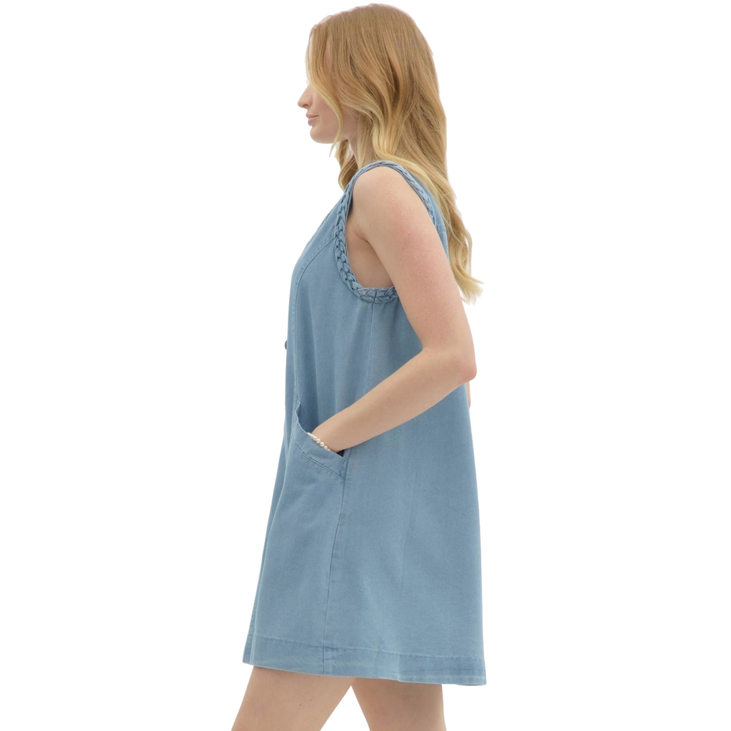 A woman is wearing a light blue denim sleeveless dress with a V-neck, buttons down the front, two large front pockets, and braided detailing on the shoulders.