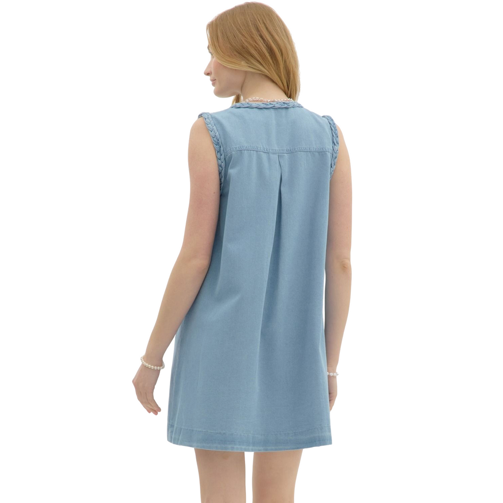 A woman is wearing a light blue denim sleeveless dress with a V-neck, buttons down the front, two large front pockets, and braided detailing on the shoulders.