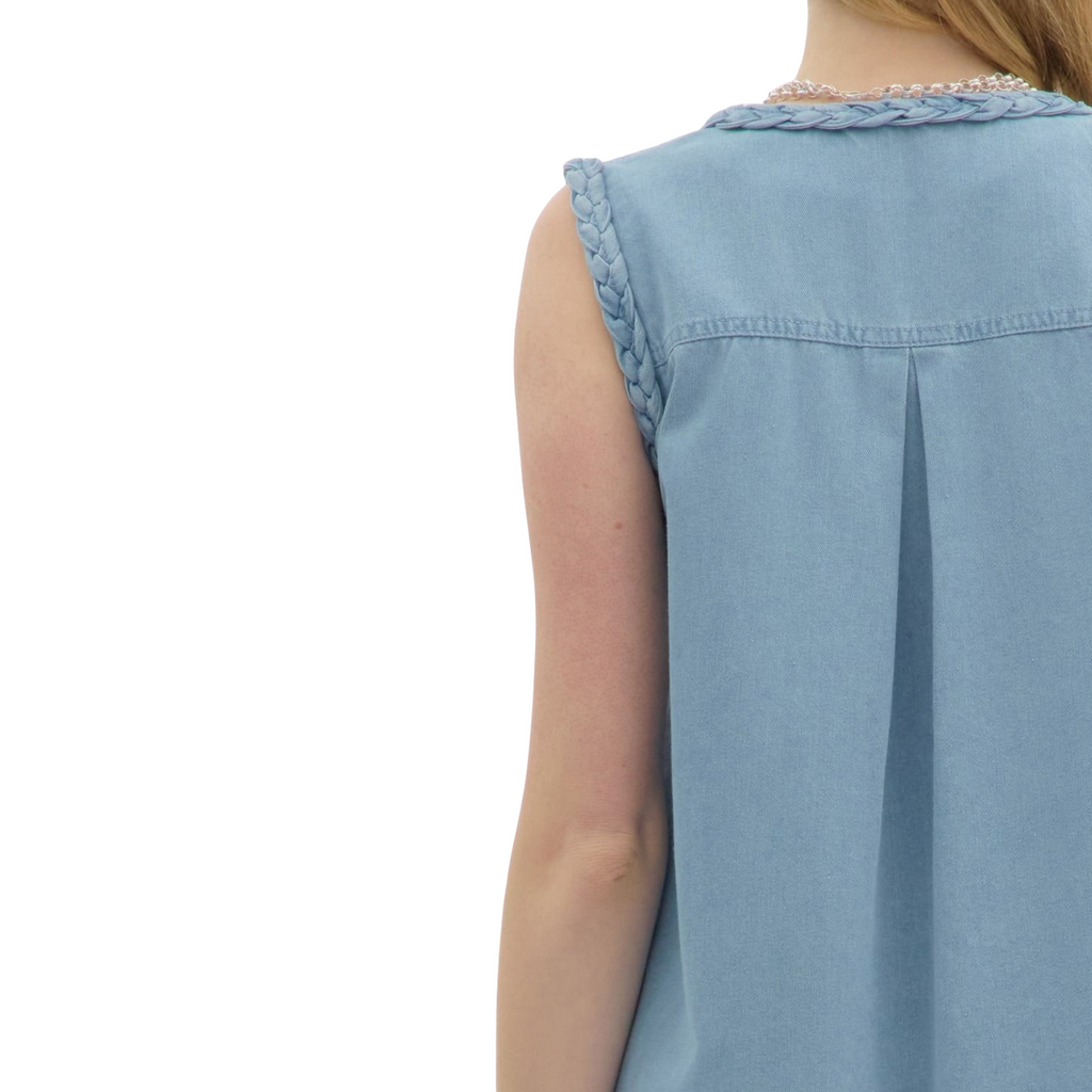 A woman is wearing a light blue denim sleeveless dress with a V-neck, buttons down the front, two large front pockets, and braided detailing on the shoulders.