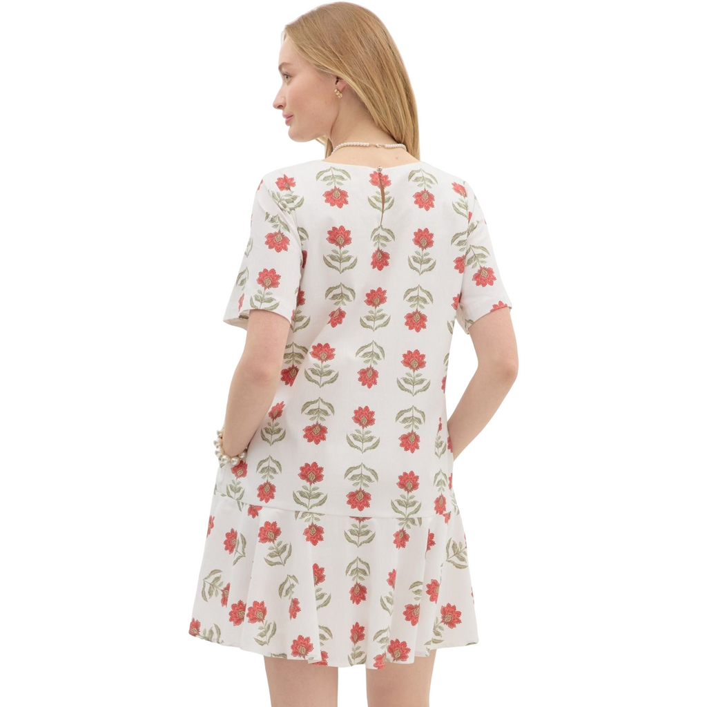 A woman is wearing a white dress with short sleeves and a ruffled hem, adorned with a repeating pattern of stylized red flowers with green stems and leaves.