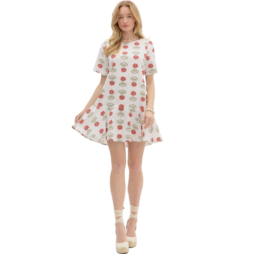 A woman is wearing a white dress with short sleeves and a ruffled hem, adorned with a repeating pattern of stylized red flowers with green stems and leaves.