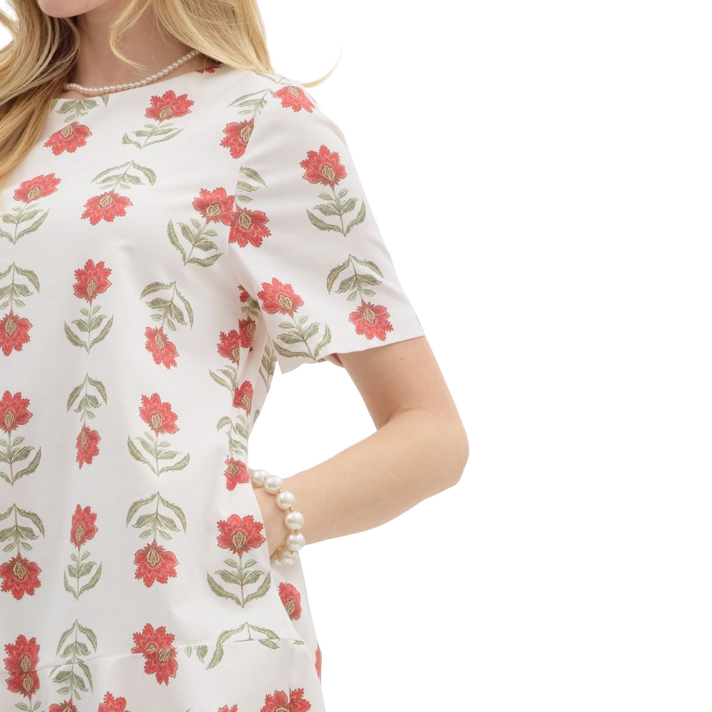 A woman is wearing a white dress with short sleeves and a ruffled hem, adorned with a repeating pattern of stylized red flowers with green stems and leaves.